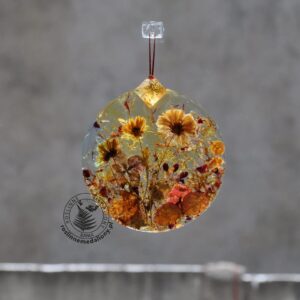Ręcznie wykonany, okrągły medalion z żywicy, w którym zatopiono różnobarwne suszone rośliny, zawieszony na szybie okna.