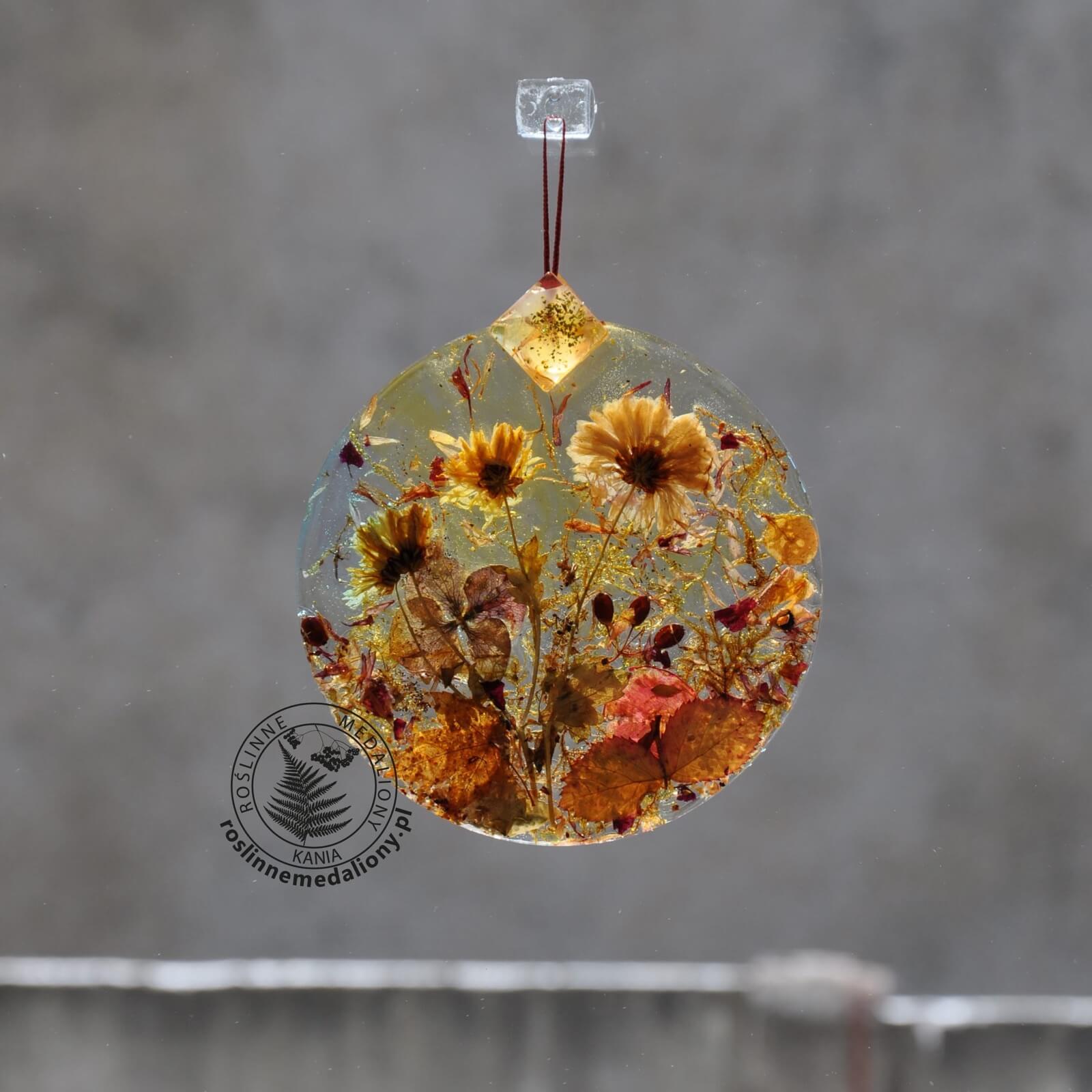 Ręcznie wykonany, okrągły medalion z żywicy, w którym zatopiono różnobarwne suszone rośliny, zawieszony na szybie okna.